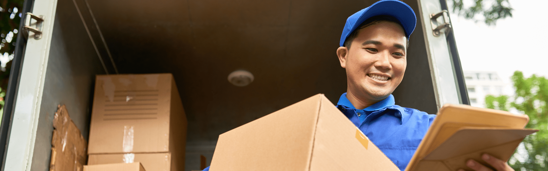 man in blue shirt and blue hat delivery uniform standing in front of delivery truck stacked with boxes while holding a box and a package