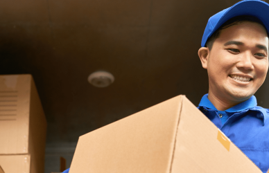 man in blue shirt and blue hat delivery uniform standing in front of delivery truck stacked with boxes while holding a box and a package
